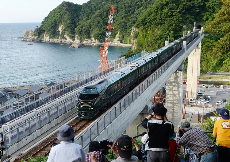 日本海をバックに橋を渡る「トワイライトエクスプレス瑞風」の1番列車と撮影する鉄道ファンたち。有名な撮影スポットとして知られる余部橋梁は空の駅として展望台などが整備されている。（時事通信フォト）