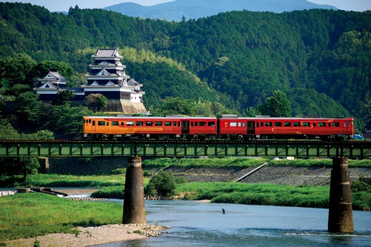 肱川の河畔に臨む大洲城近くの鉄橋を徐行する「伊予灘ものがたり」。通過時は本丸から地元の人々がのぼり旗を振って歓迎する（写真／JR四国）