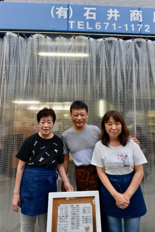 軒先で飲む客らのために雨除けカーテンをつけた店先で、3代目店主の石井康裕さん（中）、妻の理加さん（右）と母トミ子さん（左）