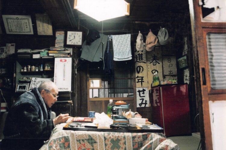 洗濯物などに囲まれて弁当を食べる老人。『ぼくは独り暮らしの老人の家に弁当を運ぶ』より