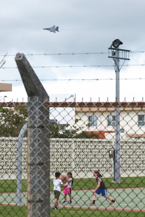 嘉手納飛行場の施設内で遊ぶ子どもたちの上空には、米軍戦闘機が飛行する。家族などの関係者も含めると、沖縄に在住する米軍関係者の数は約4万7300人（2011年時点。以後は非公表）。