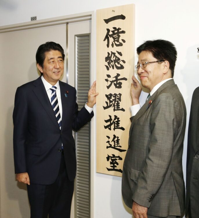 「1億総活躍推進室」の看板を掛け、笑顔の安倍晋三首相（左）と加藤勝信１億総活躍担当相（2015年10月15日当時）。推進室は2021年11月に閉鎖された（時事通信フォト）