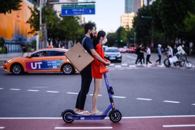 韓国、ソウルで電動キックボードに二人乗りする男女。2021年。急速に普及したもののヘルメット着用義務化に伴いシェアリングサービスでは需要が急減（AFP＝時事）