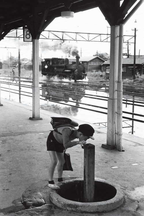 1958年、川越線大宮駅。台風で浸水した線路の手前ホームで小学生が水を飲む。写る列車は小型機関車B6（C）広田尚敬